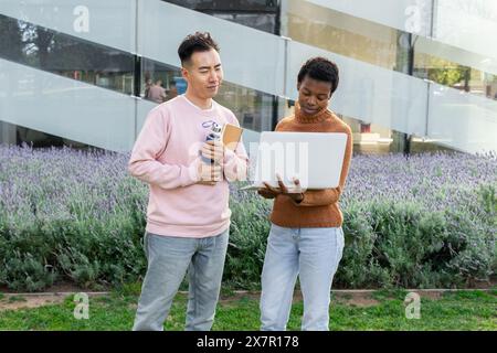 Ein asiatischer Mann und eine afrikanische Studentin studieren gemeinsam mit einem Laptop draußen. Er hält Bücher und sieht sie an, während sie f ist Stockfoto