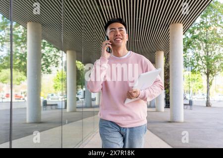 Lächelnder junger asiatischer Mann, der auf einem Smartphone spricht und draußen unter moderner Architektur ein Tablet hält. Stockfoto
