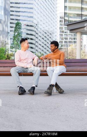 Ein junger asiatischer Mann und eine afrikanische Frau sitzen auf einer Sitzbank im Freien und unterhalten sich freundlich, umgeben von urbaner Architektur. Stockfoto