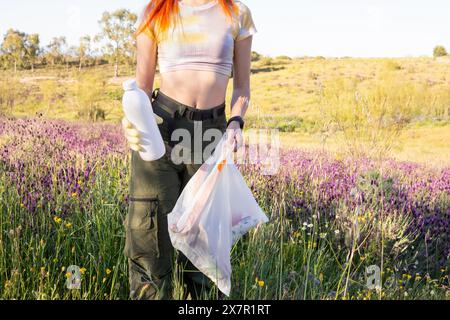 Eine junge Frau mit leuchtend orangefarbenen Haaren sammelt Müll in einem Feld aus violetten Wildblumen, trägt eine Plastikflasche und einen Müllbeutel Stockfoto