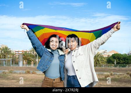 Weibliche Paare halten eine Regenbogenfahne hoch in der Luft, ein Symbol für LGBTQ-Stolz, während sie draußen mit einem hellen Lächeln posieren. Sie suchen direkt Stockfoto