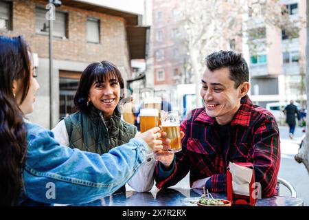 Eine Gruppe von Freunden, die einen fröhlichen Toast mit Biergläsern an einem Café-Tisch im Freien genießen und einen lebhaften Tag in der Stadt genießen. Stockfoto