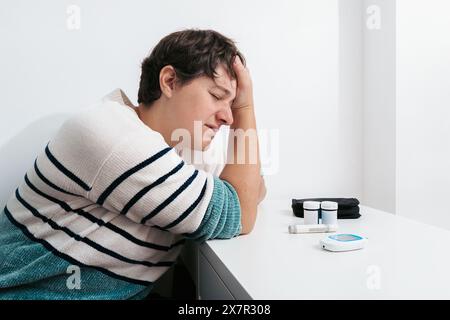 Eine Frau sitzt an einem Tisch und sieht mit der Hand am Kopf beunruhigt aus, neben Blutzuckermessgerät und Medikamenten. Sie zeigt einen Moment der Hypoglykämie Stockfoto