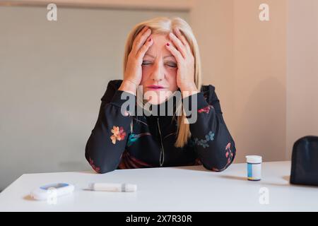 Eine Frau sitzt an einem Tisch, sichtbar beunruhigt, während sie ihren Kopf in den Händen hält. Ein Blutzuckermessgerät und Medikamente werden vor ihm sichtbar angezeigt Stockfoto