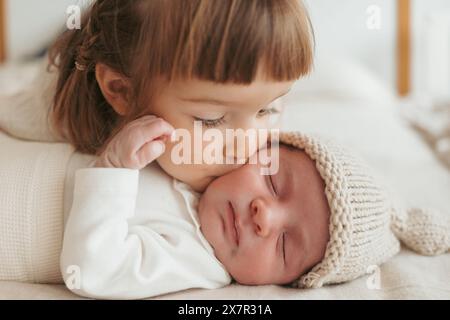 In einer herzerwärmenden Szene küsst ein Kleinkind sanft sein neugeborenes Geschwister, das mit einer gemütlichen Strickmütze schläft Stockfoto