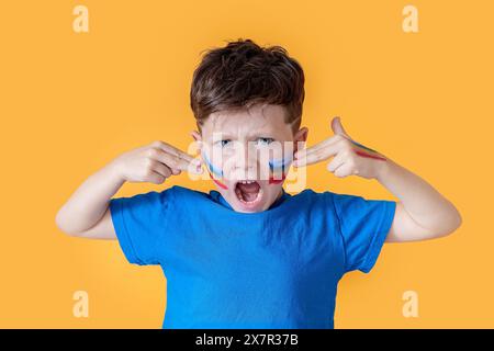 Ein kleiner Junge in einem blauen Hemd, mit Gesichtsfarbe in blauen und roten Streifen, macht eine heftige Geste vor einem hellgelben Hintergrund Stockfoto