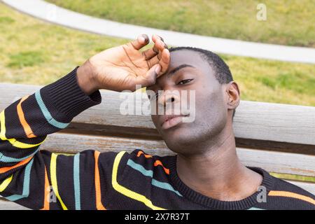 Ein junger afroamerikanischer Mann sieht müde aus und hält den Kopf, der Symptome einer Hypoglykämie veranschaulicht, und sitzt auf einer Parkbank mit grasbewachsenem Hintergrund Stockfoto