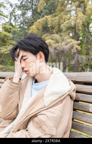 Ein junger asiatischer Mann sitzt auf einer Parkbank und drückt seine Hand auf seine Stirn. Anzeichen von Müdigkeit oder Schwindel, möglicherweise aufgrund von Hypoglykämie Stockfoto