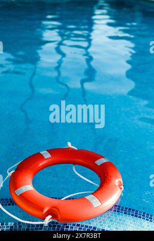 Ein roter Rettungsschirm schwimmt sanft auf dem ruhigen Wasser eines blau gefliesten Swimmingpools, reflektiert das Licht und wirft wellige Schatten auf den Poolboden Stockfoto