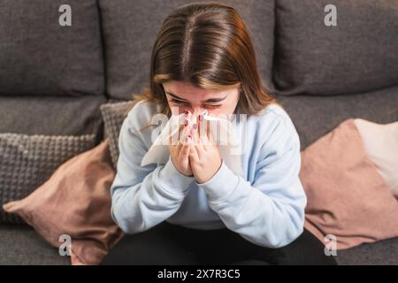 Eine junge Frau mit braunem Haar, die auf einer grauen Couch sitzt und sich mit einem Tuch die Nase bläst, möglicherweise an Erkältung oder Allergien leidet Stockfoto