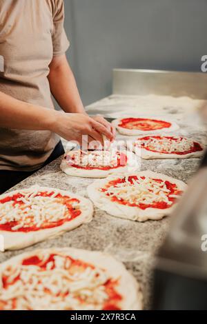 Ein anonymer Koch bastelt Pizzen, verteilt Soße und Käse auf frischem Teig, in einer kommerziellen Küche Bild fokussiert auf Hände und Pizzen, mit Facele Stockfoto