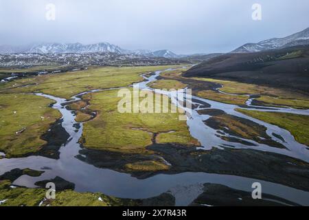 Eine ruhige Hochlandlandschaft mit einem sich windenden Fluss, der durch üppige grüne Ebenen mit schneebedeckten Bergen im Hintergrund führt Stockfoto