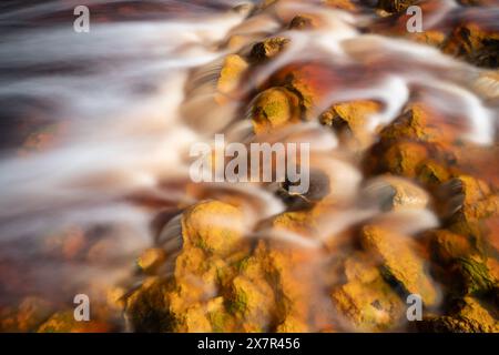 Ein Nahaufnahme-Foto zeigt den dynamischen Wasserfluss über lebendige, orange gefärbte Felsen im Riotinto River und hebt die natürliche mineralreiche Landung hervor Stockfoto