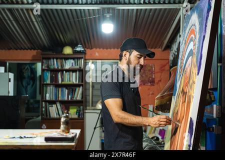 Seitenansicht eines männlichen lateinamerikanischen Künstlers malt intensiv auf einer Leinwand in seinem Atelier, Seitenansicht, während er sich auf sein Kunstwerk konzentriert. Stockfoto