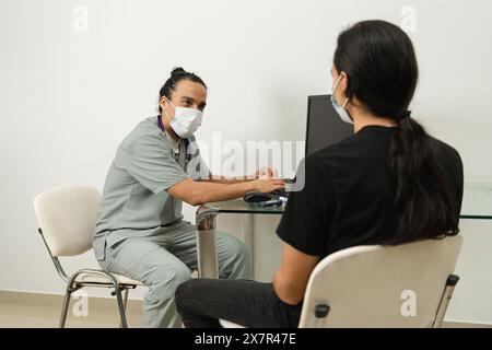Ein medizinisches Fachpersonal, das eine Gesichtsmaske trägt, diskutiert Hypoglykämie mit einer Patientin in einer klinischen Umgebung und konzentriert die Aufmerksamkeit auf einen Computer s Stockfoto