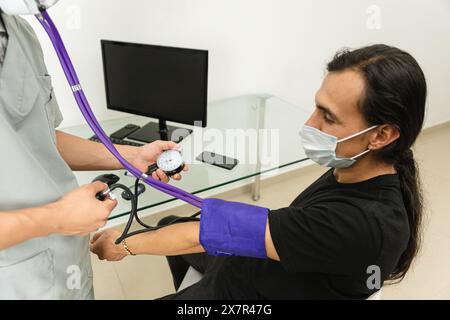 Ein medizinisches Fachpersonal im weißen Fell kontrolliert in einer modernen Arztpraxis den Blutdruck eines männlichen Patienten mit langen Haaren Stockfoto