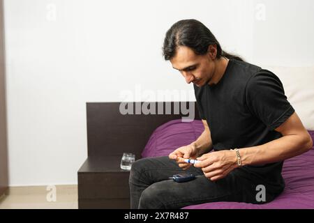 Ein erwachsener Mann sitzt auf einem Bett, kontrolliert mit einem tragbaren Messgerät seinen Blutzucker und demonstriert die routinemäßige Behandlung von Hypoglykämie in einem Haus Stockfoto