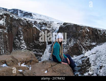 Eine junge Frau ruht auf einem Felsen mit Islands unverwechselbaren schneebedeckten Basaltsäulen und einer Bergkulisse, die in Winterkleidung gekleidet ist Stockfoto