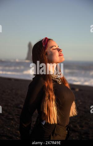Eine junge Frau sonnt sich im Sonnenlicht an einem schwarzen vulkanischen Sandstrand in Island, mit den Meereswellen und einem felsigen Meeresstapel im Hintergrund Stockfoto