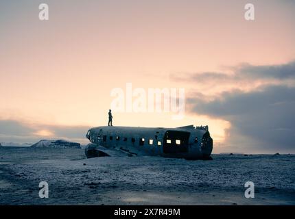 Eine Einzelfigur steht auf dem ruinierten Rumpf eines Flugzeugs, die während der Abenddämmerung vor einem pastellfarbenen Himmel in der einsamen Landschaft Islands eine Silhouette bildet Stockfoto