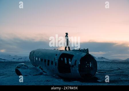 Eine Person steht auf den Überresten eines abgestürzten Flugzeugs in der verschneiten Landschaft Islands während der Dämmerung, die heitere und doch unheimliche Szene fängt ein Gefühl von Stockfoto