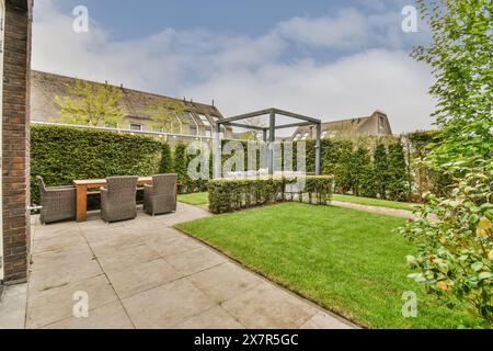 Ein üppiger Vorstadtgarten mit einem geräumigen Rasen, Der von dichten Hecken umgeben ist. Ein Essbereich mit Tisch und Stühlen ist teilweise mit modernem Metall bedeckt Stockfoto