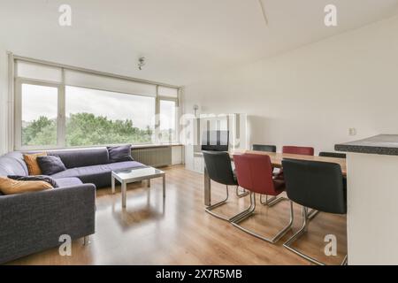 Ein geräumiges Wohnzimmer mit großem Ecksofa, Couchtisch, Essstühlen und einem breiten Fenster mit Blick auf das Grün. Stockfoto