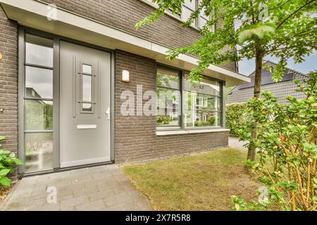 Die Fassade eines modernen Vorstadthauses mit einer stilvollen grauen Eingangstür, großen Fenstern und roten Ziegelwänden ist von Grün umgeben Stockfoto