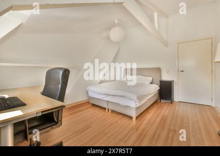 Ein gemütliches, minimalistisches Schlafzimmer im Dachgeschoss mit einem komfortablen Bett und einem kompakten Arbeitsplatz mit Laptop und schwarzem Bürostuhl. Stockfoto