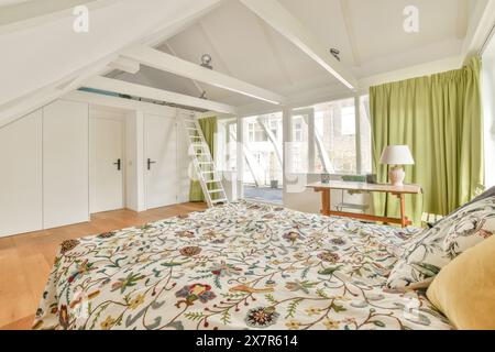 Das helle Schlafzimmer im Dachgeschoss in Schapenburgpad, Amsterdam, verfügt über weiße Wände, Balken und grüne Vorhänge. Das Zimmer verfügt über ein großes Bett mit geblümtem bedd-design Stockfoto