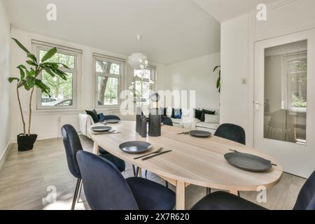 Ein gut beleuchteter, moderner Speisesaal mit einem großen Holztisch, stilvollen blauen Stühlen und viel Tageslicht durch mehrere Fenster Stockfoto