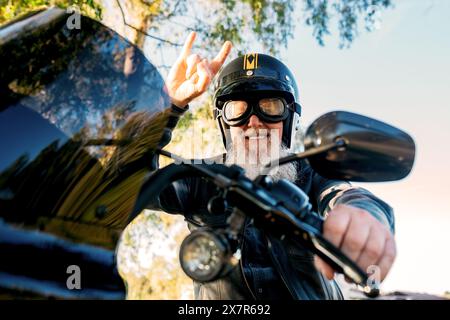 Von unten gibt ein fröhlicher Seniorenmann mit Bart und Helm ein Friedenszeichen, während er auf dem Motorrad fährt und in die Kamera blickt. Stockfoto