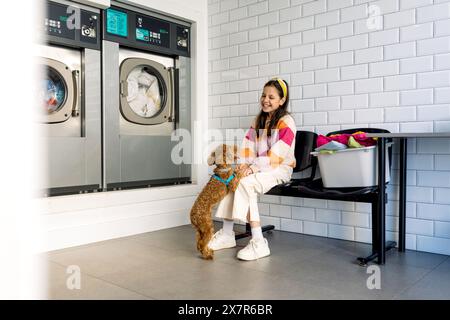 Ein lächelndes Mädchen, das in einem Wäscheraum sitzt und die Zeit mit ihrem Hund genießt, mit Waschmaschinen und einem Wäschekorb im Hintergrund. Stockfoto