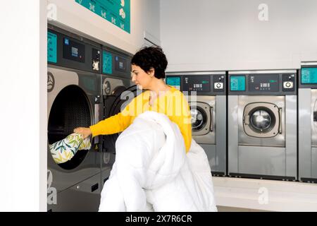 Die Seitenansicht der Frau lädt eine gemusterte Bettdecke in eine Waschmaschine in einem Waschsalon, mit mehreren Maschinen im Hintergrund Stockfoto
