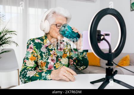 Eine ältere Frau mit einem Blumenhemd trinkt Wasser, während sie einen Podcast aufnimmt, mit Kopfhörern und einem Ringlicht in einem hellen Raum Stockfoto