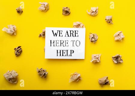 Ein gelber Hintergrund mit einem weißen Schild, auf dem steht, dass wir hier sind, um zu helfen. Das Schild ist von zerknittertem Papier umgeben, was den Eindruck eines unordentlichen oder verkrümmten Papiers erweckt Stockfoto