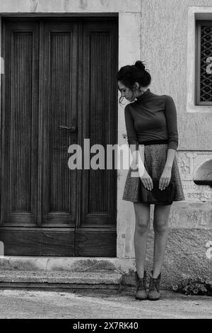 Oung Frau steht vor einer Holztür und schaut zur Seite. Sie trägt einen braunen Rollkragen und einen grauen Rock. Ihre Hände ruhen auf einer Kupplung und die s Stockfoto