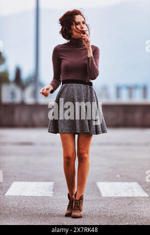 Frau, gekleidet mit braunem Rollkragen und grauem Rock, geht auf einer leeren Straße. Ihre entspannte Haltung und der ferne Blick deuten auf tiefe Gedanken hin. Der Hintergrund Stockfoto