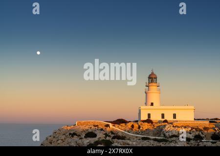 Ein vertikaler Blick auf den Leuchtturm von Cap de Cavalleria auf Menorca bei Sonnenuntergang bei aufsteigendem Vollmond Stockfoto