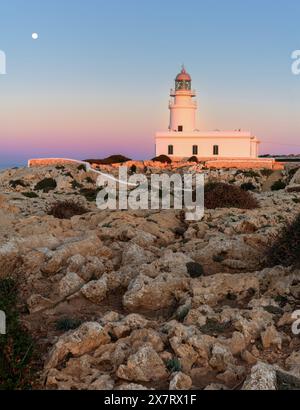 Ein vertikaler Blick auf den Leuchtturm von Cap de Cavalleria auf Menorca bei Sonnenuntergang bei aufsteigendem Vollmond Stockfoto