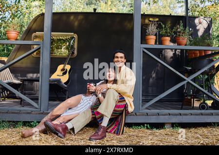 Ein interrassisches Paar sitzt auf dem Boden vor einem Wohnwagen und genießt einen romantischen Kurzurlaub in einer ruhigen natürlichen Umgebung. Stockfoto