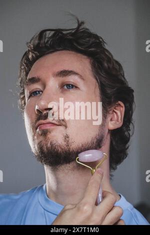 Schöner junger Mann, der sein Gesicht mit einer Rosenquarzwalze massiert Stockfoto