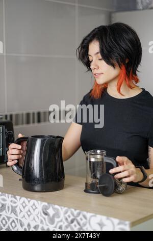 Schwarzhaarige weiße Person lächelt beim Kaffeezubereiten. Sie haben einen Wasserkocher und es gibt eine französische Presse mit gemahlenem Kaffee. Stockfoto