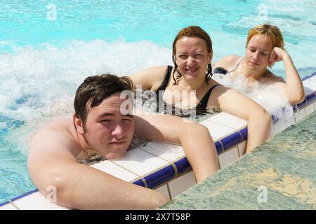 Eine vielfältige Gruppe von Menschen, darunter Familien und Freunde, wird in einem lebendigen Swimmingpool mit Hydromassagedüsen schwimmen, spielen und entspannen sehen. Stockfoto