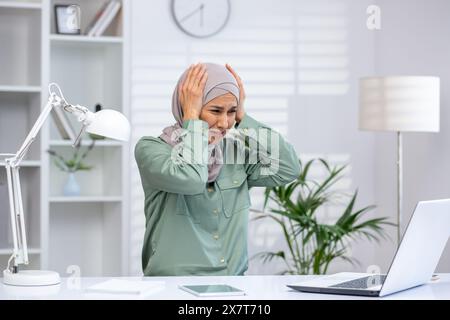 Eine gestresste Geschäftsfrau mit Hijab, die an ihrem Schreibtisch mit einem Laptop arbeitet und einen überwältigenden Ausdruck zeigt. Das Konzept von Stress, Frustration und Arbeitsdruck. Stockfoto