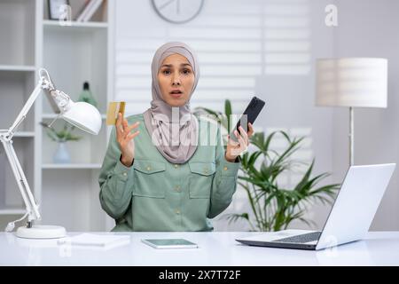 Schockierte junge Frau mit Hijab, Telefon und Kreditkarte in einem modernen Büro mit Laptop. Stockfoto