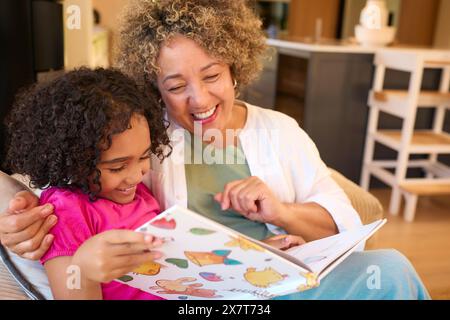 Großmutter kümmert sich um Enkelin zu Hause, sitzt auf Sofa und liest gemeinsam Eine Geschichte Stockfoto