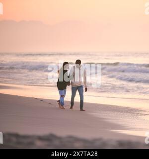Lässig Gekleidetes, Liebevolles Junges Paar, Das Am Sunrise Entlang Der Strandküste Geht Und Hände Hält Stockfoto