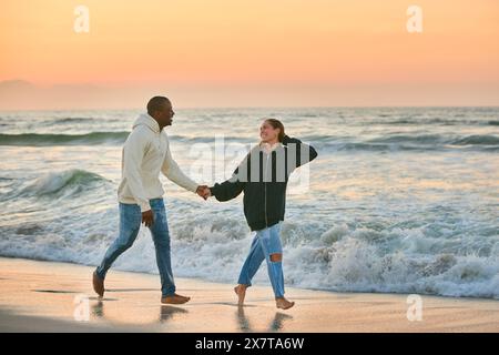 Lässig Gekleidetes, Liebevolles Junges Paar, Das Bei Sonnenaufgang Entlang Der Beach Shoreline Durch Die Wellen Läuft Stockfoto
