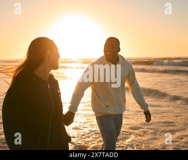 Lässig Gekleidetes, Liebevolles Junges Paar, Das Bei Sonnenaufgang Entlang Der Beach Shoreline Durch Die Wellen Läuft Stockfoto